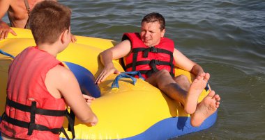 Kırmızı can yelekli baba ve oğlu şişirilebilir sarı bir tüple denizde birlikte seyahat ediyorlar. Ekstrem Spor. Adrenalin. Yaz eğlencesi ve etkileyici hız pateni. Aktif yaşam tarzı. Mutlu aile kavramı