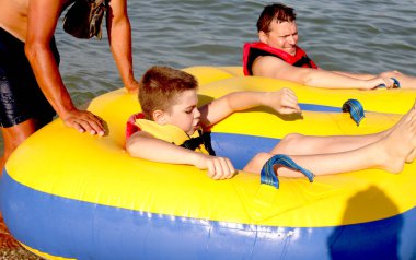 Kırmızı can yelekli baba ve oğlu şişirilebilir sarı bir tüple denizde birlikte seyahat ediyorlar. Ekstrem Spor. Adrenalin. Yaz eğlencesi ve etkileyici hız pateni. Aktif yaşam tarzı. Mutlu aile kavramı