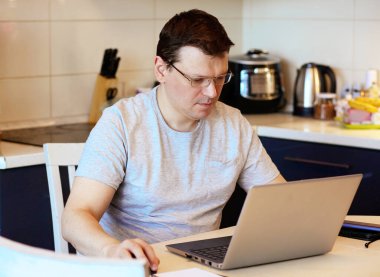 2019-Ncov salgını sırasında evde çalışmak. Odaklanmış ve ciddi gözlüklü genç adam evde mutfakta otururken dizüstü bilgisayarla çalışıyor. Koronavirüs salgını sırasında işyerinde. Yalıtım