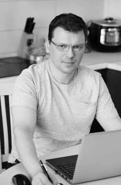 Work at home online during a pandemic 2019-ncov. Focused and serious young man in glasses working on laptop while sitting in the kitchen at home. Workplace during the coronavirus pandemic. Insulation