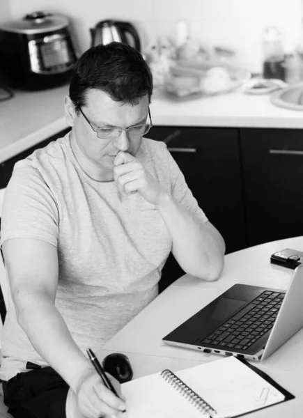 Work at home online during a pandemic 2019-ncov. Focused and serious young man in glasses working on laptop while sitting in the kitchen at home. Workplace during the coronavirus pandemic. Insulation