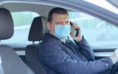 Koruyucu maskeli genç bir adam soldan çekişli bir arabada oturuyor ve cep telefonundan kameraya bakıyor. Tıbbi maskeli bir şoför. Coronavirus covid-19, salgın. Virüs koruması