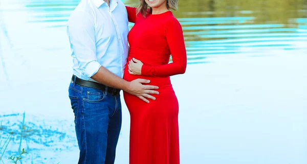 Married couple. Pregnant womanin a red dress and man in a white shirt and jeans on the lake. Love, family, pregnancy, motherhood. The concept of a happy marriage and family. Copy space.