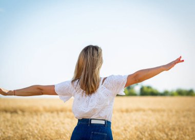Beyaz bluz ve buğday tarlasında mavi kot etek giyen sarışın, beyaz ve mutlu kız özgürlüğün tadını çıkarıyor. İnsanları doğayla uyum içinde kavrayın. Kadın psikolojisi, duyguları ve duyguları.