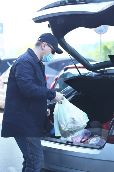A man in a protective mask, a light windbreaker and black glasses loads food into the trunk of a car. Essential goods. The life of urban residents. Coronavirus and pandemic concept. COVID-19