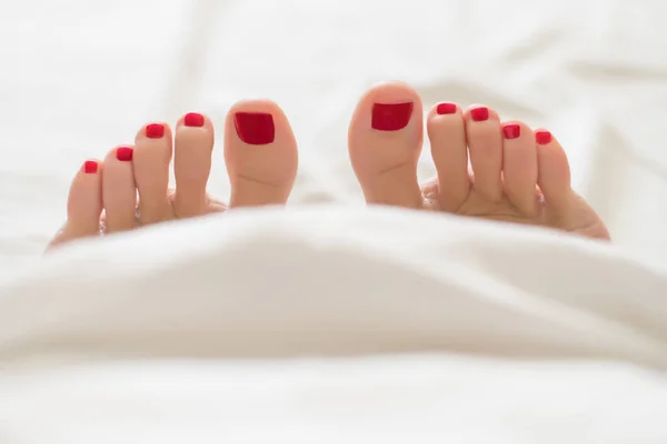 Dedo femenino con pedicura roja en la cama de debajo de las cubiertas blancas Primer plano de los pies femeninos cubiertos con pedicura roja — Foto de Stock
