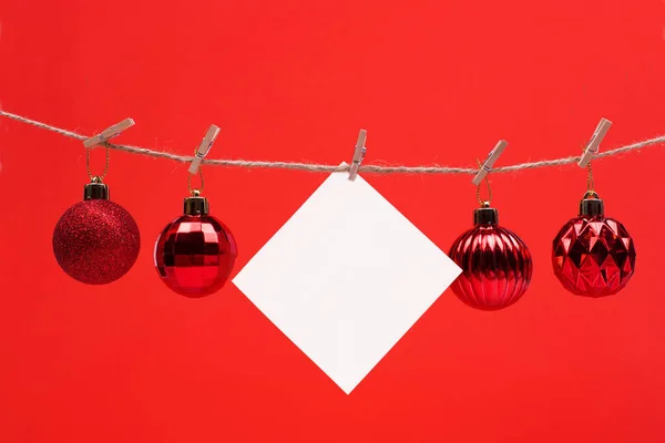Een wit vierkant velletje nota papier hangt op wasknijpers op een rode achtergrond met kerstbal, mock up — Stockfoto