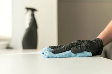 Siyah eldivenli bir kadının elinde mavi bir mikrofiber temizleme bezi ve sterilize edici solüsyonla dezenfeksiyon ve temizlik malzemesi bulunur..