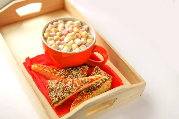Cocoa drink with colorful small marshmallows and pieces of sweet homemade cake in a wooden tray.