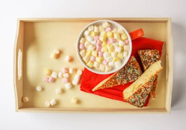 Cocoa drink with colorful small marshmallows and pieces of sweet homemade cake in a wooden tray.