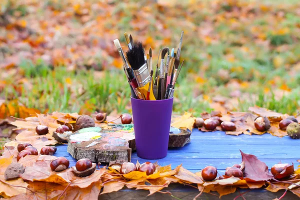 Workplace for painter in autumn park with wood palette and paintbrushes in plastic container. Fall leaves and chestnuts on blue wooden background.