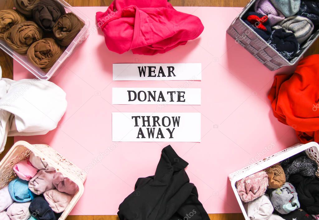 Clothes sorting in home wardrobe for donation, wearing and discard with paper notes on soft pink background. Clothing items in textile and plastic boxes.