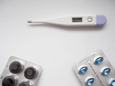 Tablet ve kapsüller şeklinde ilaçlar yuvarlak ve oval mavi beyaz turuncu ve yeşil renkli açık arkaplanda elektronik beyaz termometreyle