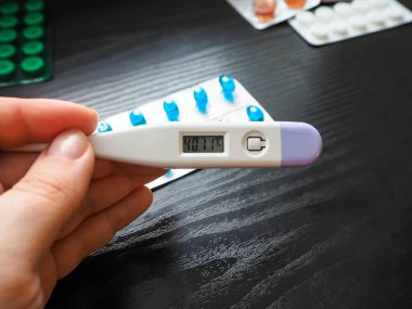 Bir kadının elinde, arka planda çok yüksek sıcaklıkta, yeşil ve mavi tabletlerle dolu beyaz bir elektronik termometre ve koyu bir arka planda duran bir çift beyaz lateks eldiven bulunur.