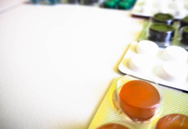 Tablet ve kapsül biçimindeki ilaçlar yuvarlak ve oval bir şeklin sağ tarafında açık arkaplanda mavi, beyaz, turuncu ve yeşil renkte sergilenmektedir.