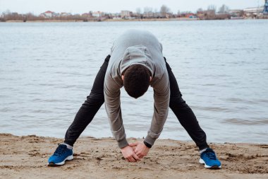 Genç yakışıklı sporcu sahilde koşmadan önce esneme hareketleri yapıyor. Koşucu sabahın erken saatlerinde sahilde egzersiz yapıyor.