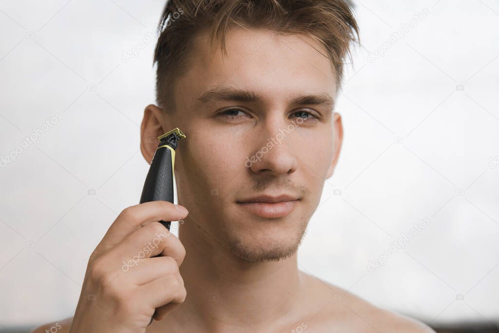 Close-up Portrait of handsome man shaving his beard with electric shaver in morning, against grey background