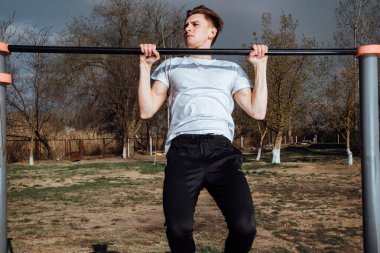 Beyaz tişörtlü genç bir adam spor sahasındaki yatay bir bara yanaşıyor. Port, sokak sporu konsepti