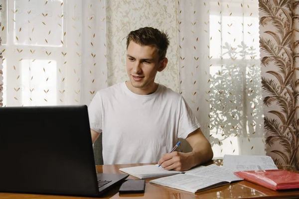 A young man does his homework in a notebook and works on a laptop at home at a Desk.A young happy male student is studying distance learning at home.Distance learning in quarantine