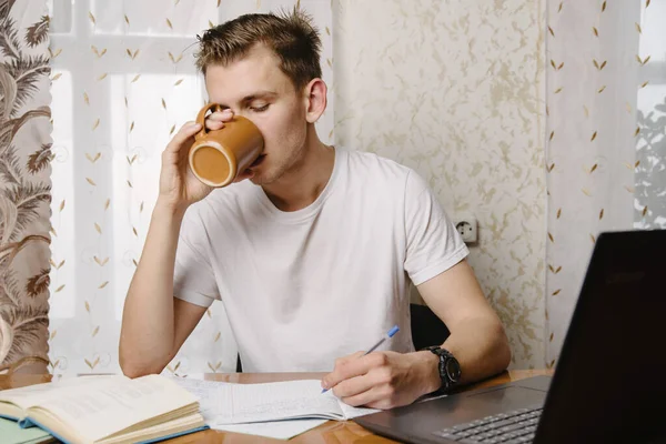 A young man does his homework in a notebook and works on a laptop at home at a Desk.A young happy male student is studying distance learning at home.Distance learning in quarantine