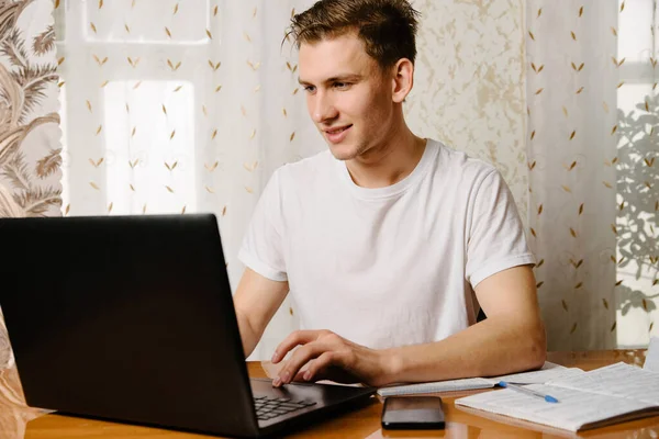 A young man does his homework in a notebook and works on a laptop at home at a Desk.A young happy male student is studying distance learning at home.Distance learning in quarantine