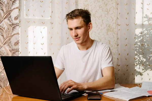 A young man does his homework in a notebook and works on a laptop at home at a Desk.A young happy male student is studying distance learning at home.Distance learning in quarantine