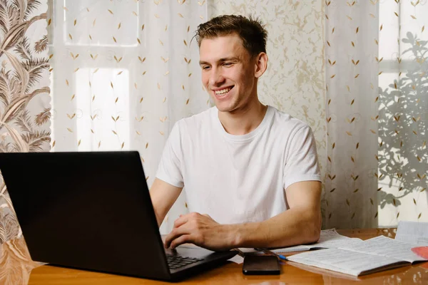 A young man does his homework in a notebook and works on a laptop at home at a Desk.A young happy male student is studying distance learning at home.Distance learning in quarantine