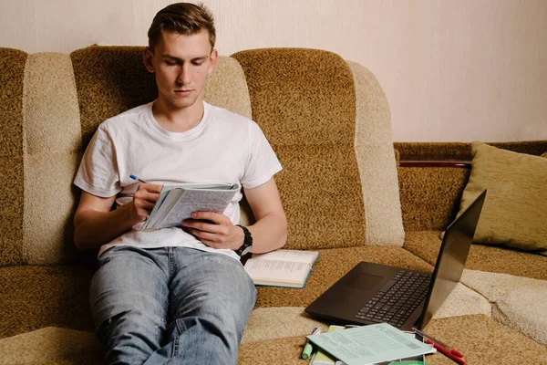A young man does his homework in a notebook and works on a laptop on the couch.A happy male student is studying distance learning at home.Online learning in quarantine