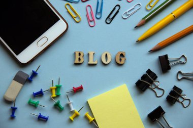 Kağıt ataç, kalem, kalem, kağıt, klasör, silgi, düğme, cep telefonu etrafında tahta harflerden oluşan blog kelimesi. Yaratıcı bir kavram. Üst manzara, düz uzanma.