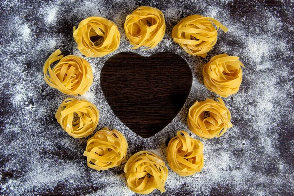 Raw round pasta, sprinkled flour on a dark background. In the center is a heart shape. The concept of delicious food. Top view, flat lay, copy space.