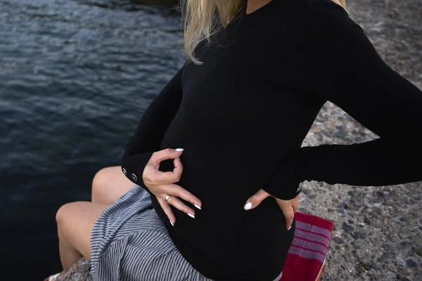 Pregnant woman sits near the sea and shows that she loves her ch — Stock Photo, Image