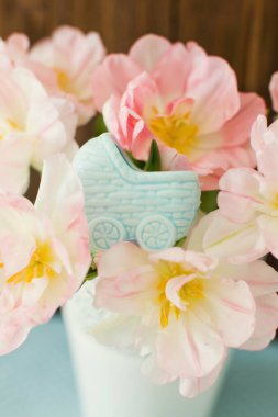 Mavi bebek arabası heykelciği, bebeği bekliyor. Hamilelik teması. Beyaz metal vazoda pembe ve beyaz taç yapraklı lale demeti.