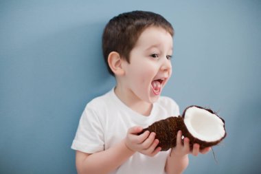 Beyaz tişörtlü ve sarı şortlu küçük çocuk mavi duvarın arkasında iki hindistan cevizi tutuyor.