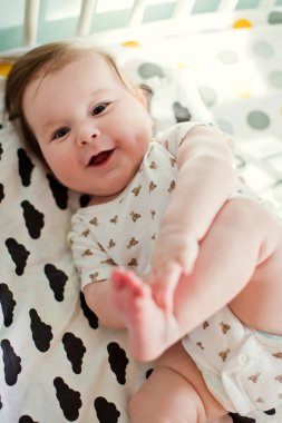 Sevimli erkek bebek yatakta uzanıyor ve bacaklarıyla oynuyor. Bebek odasında güneşli bir sabah.