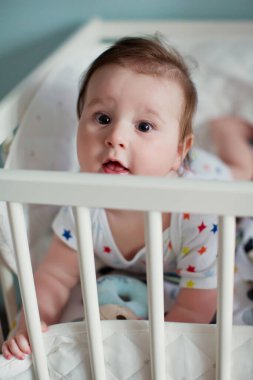 Küçük oğlan ışık odasında beşiğinde yatıyor ve oyuncaklarıyla oynuyor.