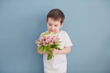 Beyaz tişörtlü küçük çocuk pembe çiçek buketi tutuyor. Mavi duvarın arkasındaki komik çocuk.