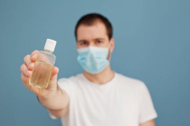 Ameliyat maskesi takan genç adam mavi arka plandaki mikroplardan ellerini temizlemek için el dezenfektanı kullanıyor.