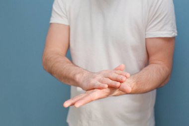 Ameliyat maskesi takan genç adam mavi arka plandaki mikroplardan ellerini temizlemek için el dezenfektanı kullanıyor.