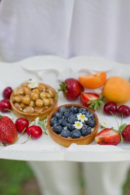 Beyaz elbiseli kadın elinde taze meyveli beyaz ahşap tepsi, böğürtlenli turtalar ve fındıklı turtalar tutuyor..