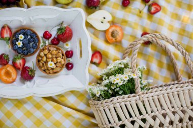 Parkta piknik. Beyaz ahşap tepside taze meyve ve tartlar, sarı masa örtüsünün üzerinde beyaz papatyalar olan saman torbası.