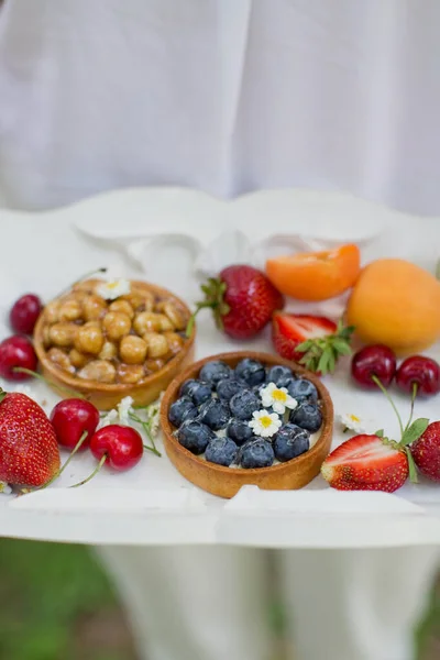Woman in a white clothes holds white wooden tray with fresh fruits and tarts with berries and nuts.