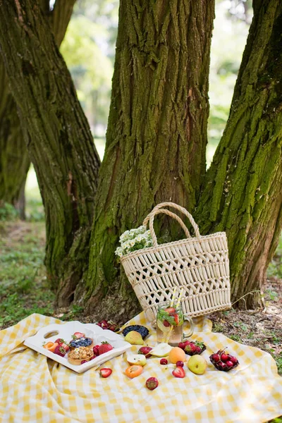 Picnic at the park. Fresh fruits and tarts on a white wooden tray, straw bag with white chamomiles near tree