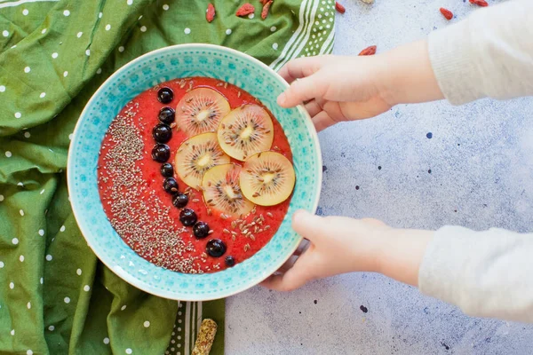 Smoothie bowl with kiwi, black berries and chia seeds and flax seeds on a light background. Child touch the bowl with smoothie