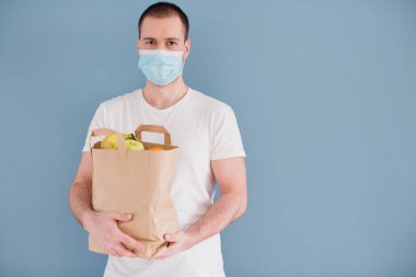 Mavi arka planda meyve ve sebzeli cerrahi maske takan beyaz bir adam. Teslimatçı.
