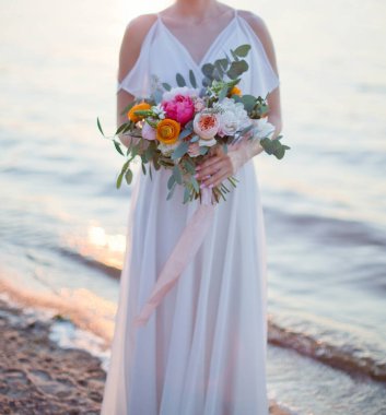 Gelin elinde güzel çiçekler ve pembe ipek kurdelelerle düğün buketi tutuyor. Deniz kenarında duran çiçekli genç bir kadın.