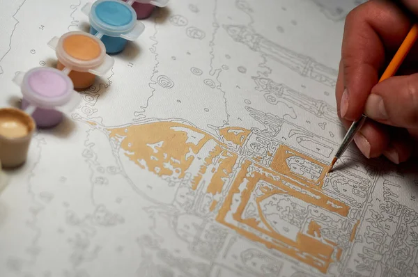 Kız eli, tuvaldeki numaralarla fırça boyasıyla resim yapıyor. Karantina sırasında yaratıcı bir hobi. Rakamlara göre resim. Tuvale resim çiziyorum. Yakın çekim, seçici odaklanma