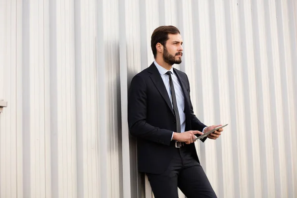 Homem elegante de pé contra uma parede cinza com um tablet — Fotografia de Stock