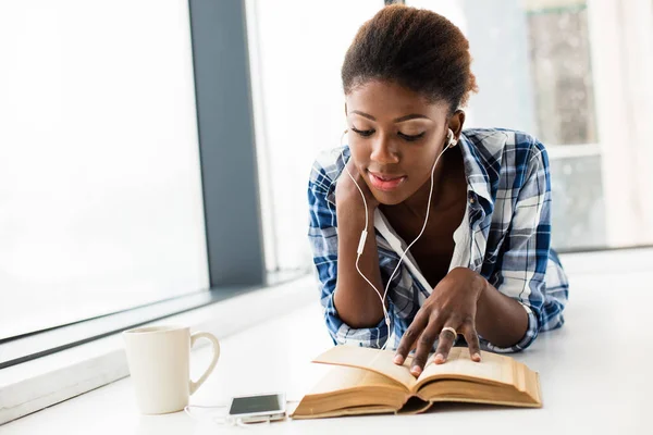 Mujer negra leyendo un libro al lado de una gran ventana con labio natural — Foto de Stock