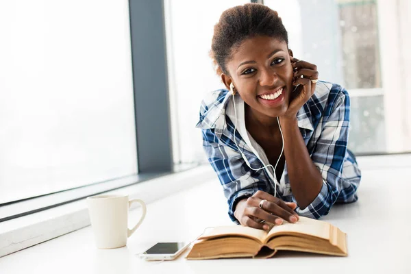 Mujer negra leyendo un libro al lado de una gran ventana con labio natural — Foto de Stock