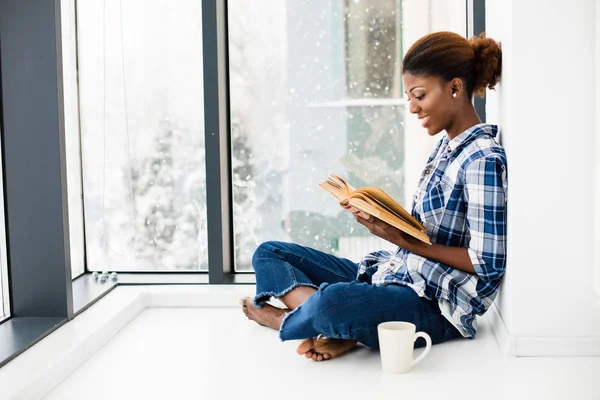 Mujer negra leyendo un libro al lado de una gran ventana con labio natural — Foto de Stock